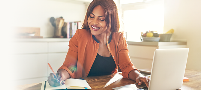 Woman writes in a notebook while looking at something on her laptop.